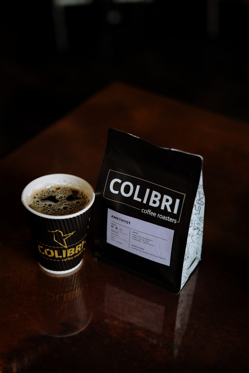A Colibri to-go cup of black coffee next to a bag of Amethyst on a dark wood table