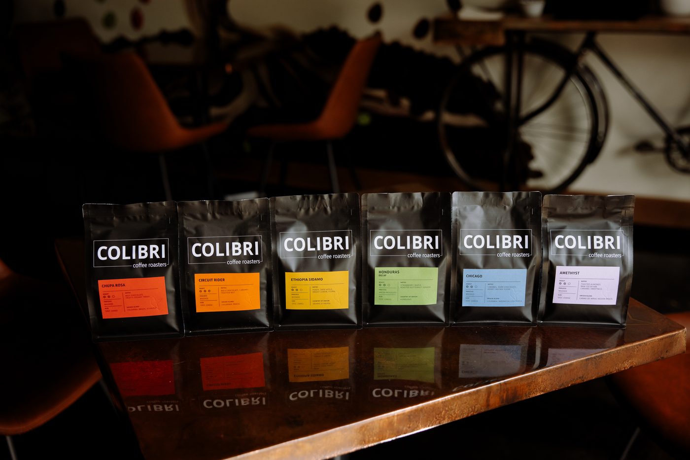 All six Colibri roasts side by side on a copper-edged table — Chupa Rosa, Circuit Rider, Ethiopia Sidamo, Honduras Decaf, Chicago and Amethyst