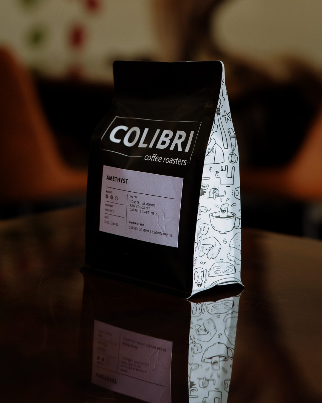 A bag of Amethyst — black Colibri bag with a lavender label and the doodle-printed side gusset, on a glossy table
