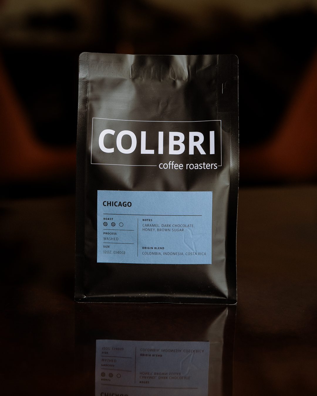 A bag of Chicago — black Colibri bag with a sky-blue label, on a glossy table in front of the coffee-tree mural