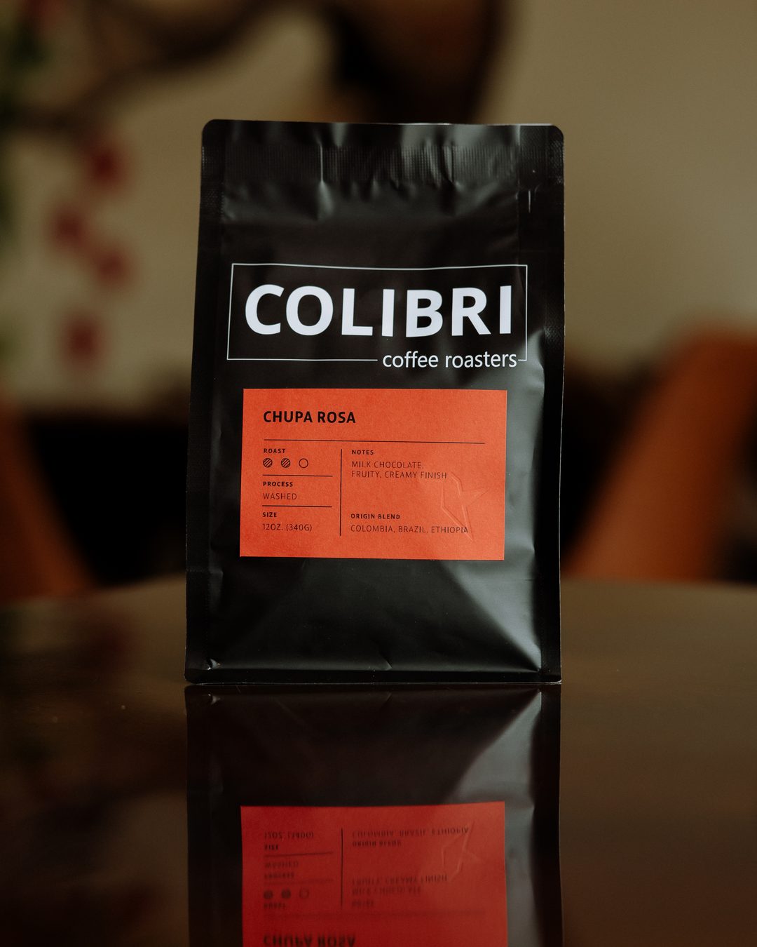 A bag of Chupa Rosa — black Colibri bag with a coral-red label, standing on a glossy table in front of the coffee-tree mural