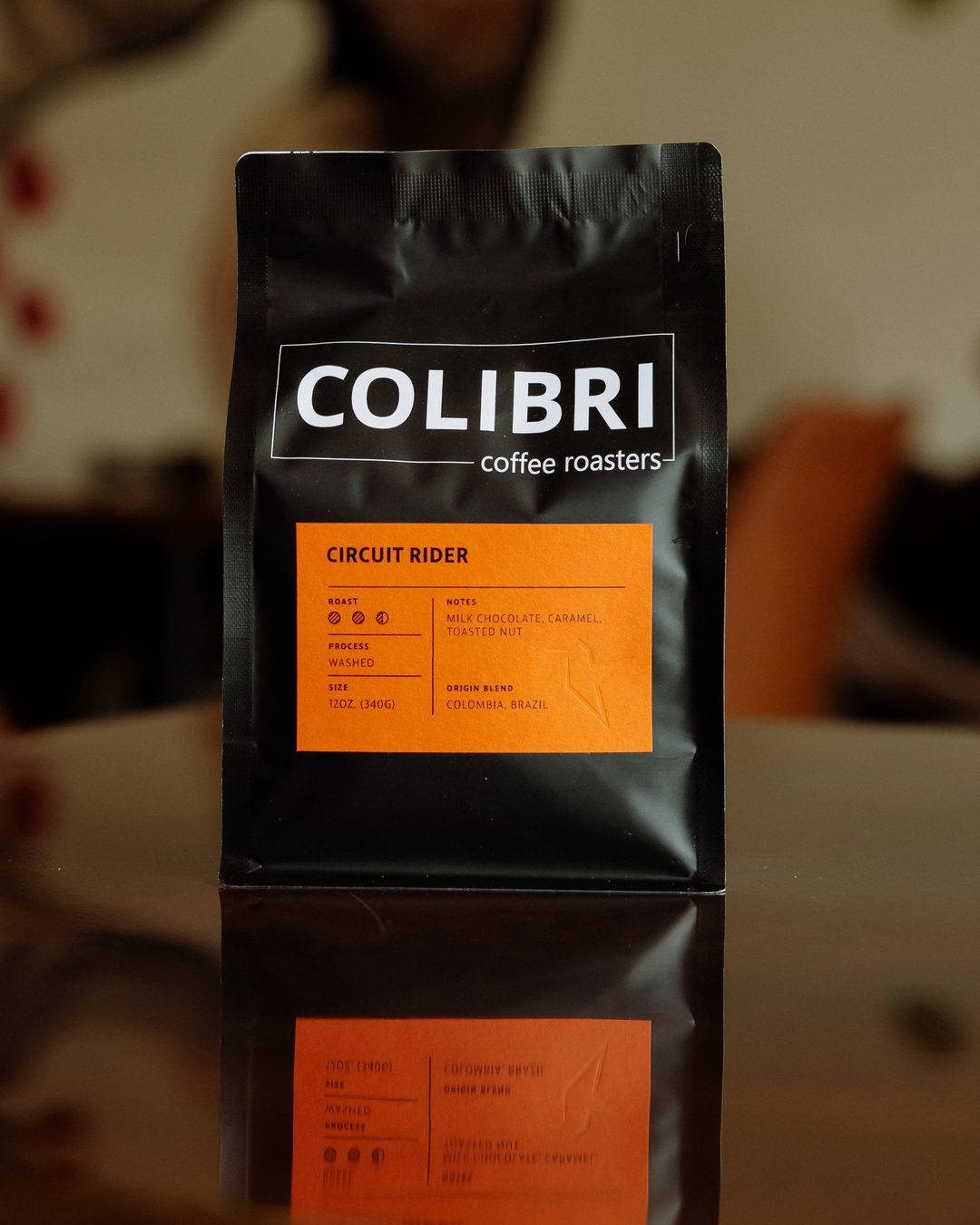 A bag of Circuit Rider — black Colibri bag with an orange label, on a glossy table in front of the coffee-tree mural