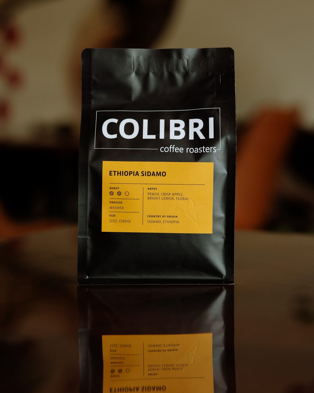 A bag of Ethiopia Sidamo — black Colibri bag with a golden-yellow label, on a glossy table in front of the coffee-tree mural