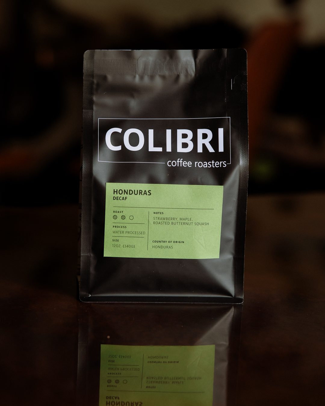 A bag of Honduras Decaf — black Colibri bag with a green label, on a glossy table in front of the coffee-tree mural