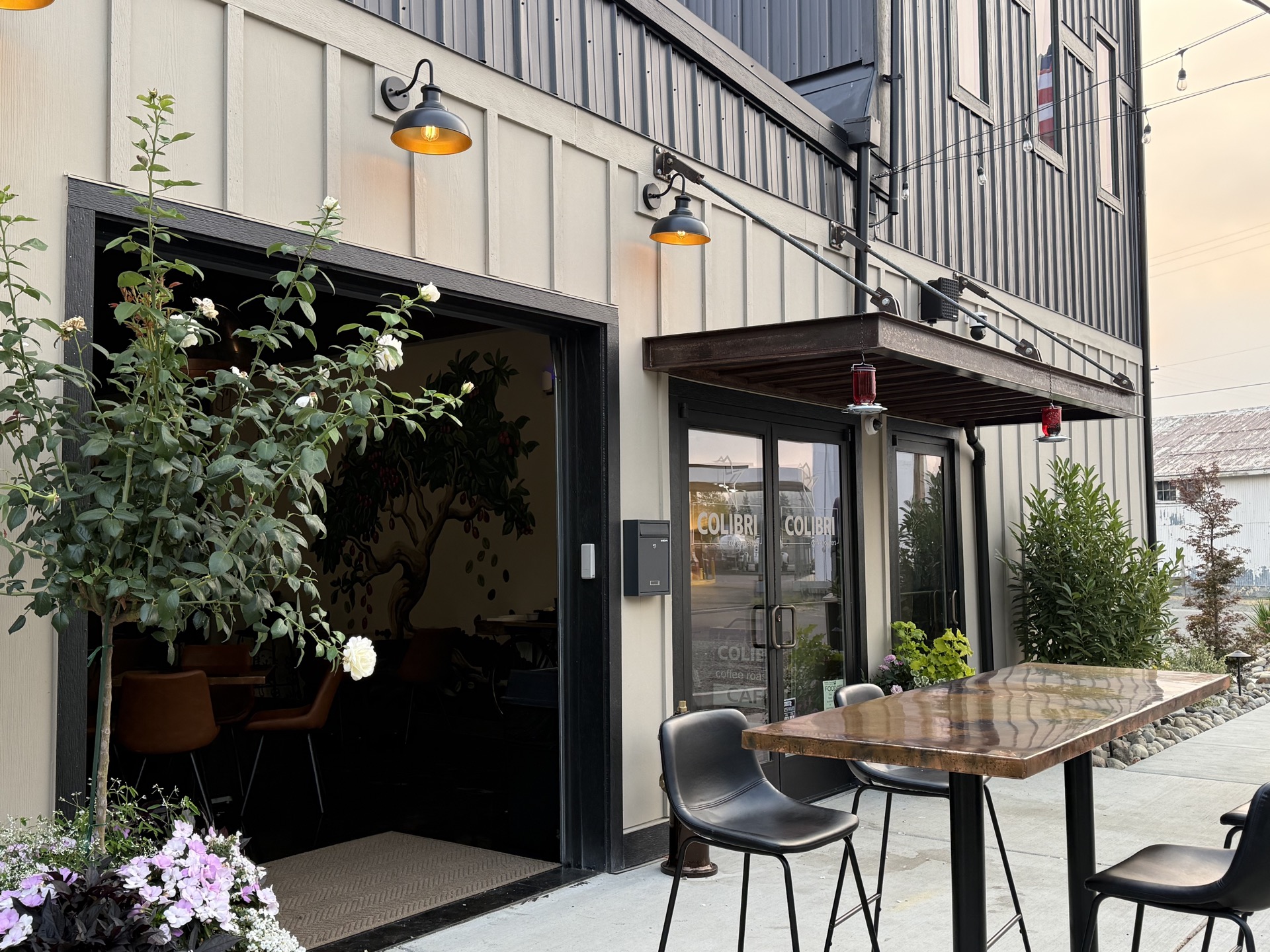 Golden hour on the Colibri patio — cream board-and-batten siding, black barn lights, the rusted-copper awning and red hummingbird feeders over the door