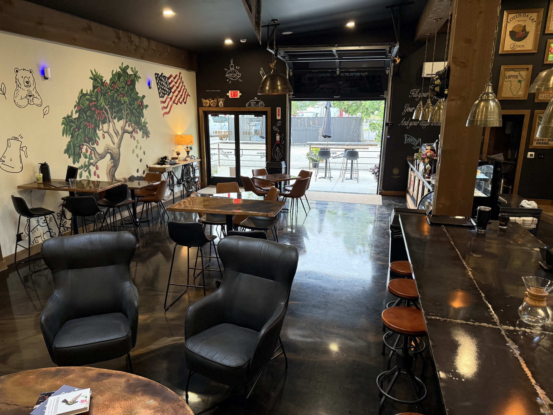 The full room — glossy dark floor, coffee-tree mural, copper tables, leather seating, and the roll-up garage door open to the street