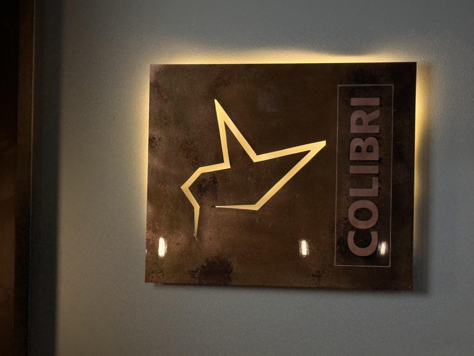 The Colibri sign — a rusted steel plate with the origami hummingbird cut out, backlit in warm gold